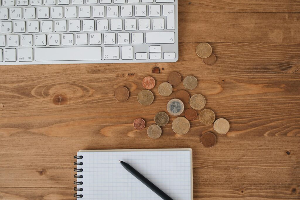 Scattered coins on a wooden desk alongside a keyboard and notebook, captured from above.